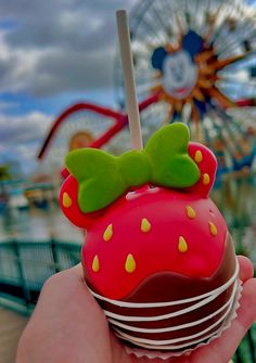a hand holding a chocolate covered strawberry with a green bow on top and a ferris wheel in the background