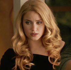 a woman with long blonde hair wearing a black dress and looking at the camera while standing in front of a window