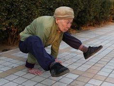 an old man is playing with his shoes on the sidewalk in front of some bushes