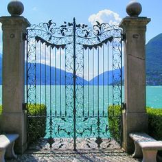 an iron gate with two stone benches in front of it and the water behind it