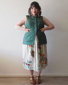 a woman standing in front of a white wall wearing a green vest and floral skirt