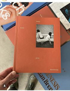 a person holding an orange book in front of several books on the ground with one hand