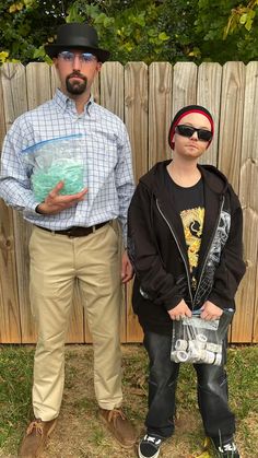 two people standing next to each other in front of a wooden fence and holding bags
