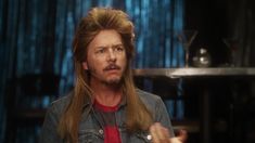 a man with long hair wearing a denim jacket and red shirt is talking to someone