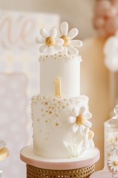a three tiered cake decorated with daisies and gold accents on a table at a baby's first birthday party