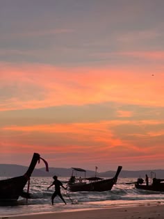 two boats on the beach at sunset with people walking in the water and one person carrying a surfboard