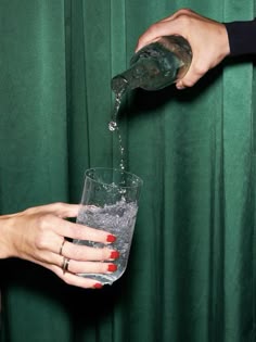 two people pouring water into a glass with red nail polishes on their hands and green curtains behind them