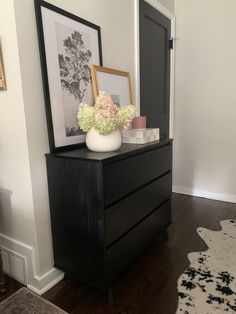 a black dresser with flowers and pictures on it in a white walled living room area