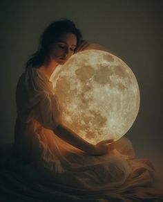 a woman sitting on the ground holding a large illuminated moon in her hands, with dark background