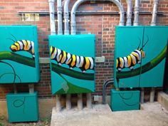 two paintings of caterpillars painted on green boxes in front of a brick wall
