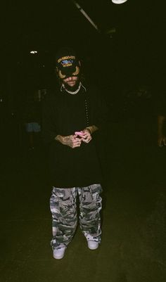 a man standing in the dark looking at his cell phone while wearing camo pants