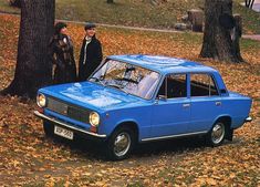 two people standing next to an old blue car in the park with fallen leaves on the ground