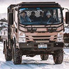 three men sitting in the drivers seat of a large truck on a snowy road with mountains in the background