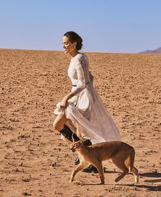 a woman in a white dress is walking with a dog on a desert plain,