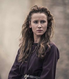 a woman with long hair standing in front of a building wearing a purple shirt and brown belt
