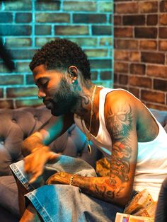 a man with tattoos sitting on a couch in front of a brick wall and looking at his cell phone