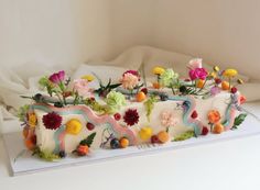 a cake decorated with flowers and fruit on a white tableclothed surface in front of a window