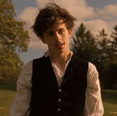 a man wearing a vest and white shirt standing in front of a field with trees