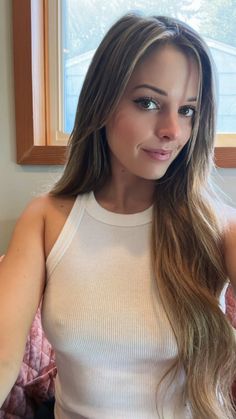 a woman with long brown hair wearing a white top and posing for the camera in front of a window