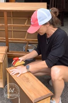 a woman in black shirt and red hat working on wooden furniture with scissors, sanding block