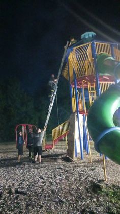 two children playing on a playground at night with the lights on and green slide in the foreground