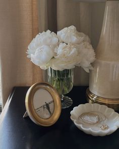 white flowers are in a vase on a black table next to a clock and lamp