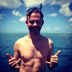 a man with no shirt on standing in front of the ocean giving two thumbs up