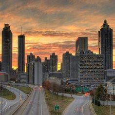 the city skyline is lit up at sunset as traffic travels down an empty highway in front of tall buildings
