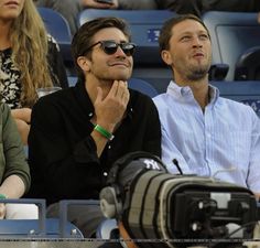 two men and a woman sitting in the stands at a tennis game, one with sunglasses on his face