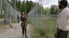two people walking down a dirt road next to a chain link fence with barbed wire