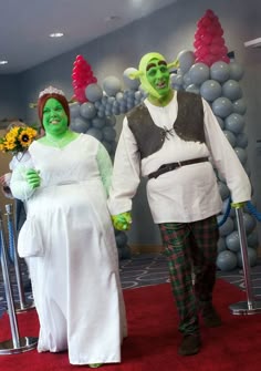 two people dressed in costumes standing on a red carpet with balloons and decorations behind them