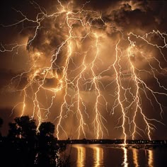 lightning strikes across the sky over water and trees in front of dark clouds with bright lights