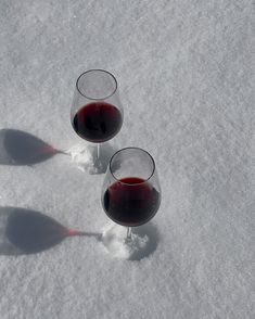 two glasses of red wine are sitting in the snow with their shadow on the ground