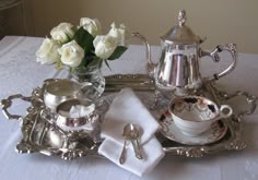 a silver tea set on a white tablecloth with flowers in a vase next to it