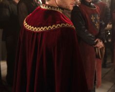 a man wearing a red cape and gold trim standing next to other men in medieval clothing