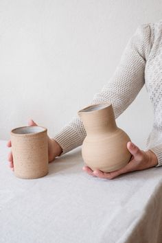 a woman is holding a vase in her right hand and another one on the table behind her