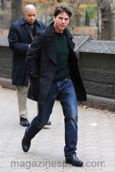 a man in black coat and jeans walking down the street with another man behind him