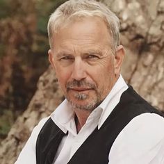 a man with grey hair and beard wearing a black vest standing in front of a tree