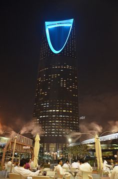 people sitting at tables in front of a tall building with a blue light on it