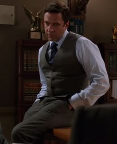 a man in a vest and tie sitting on a table next to a bookshelf