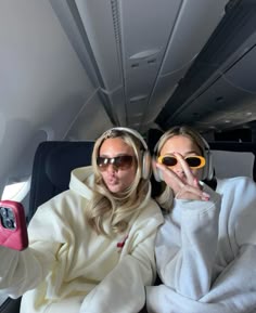 two women sitting in the back of an airplane with headphones on and one holding up her phone