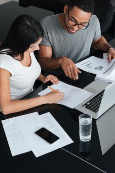 a man and woman sitting at a table working on paperwork with a laptop in front of them