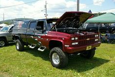 a red truck with its hood open parked in the grass next to other cars and people