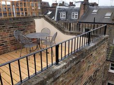 an outdoor table and chairs on a balcony overlooking the rooftops with brick buildings in the background
