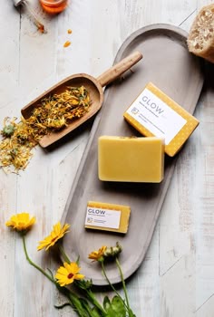 some soaps and flowers on a tray next to a wooden spoon with yellow flowers