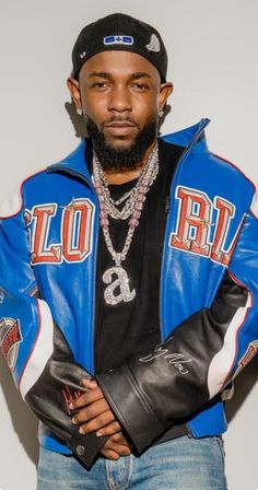 a man with a beard wearing a blue leather jacket and black hat standing in front of a white wall