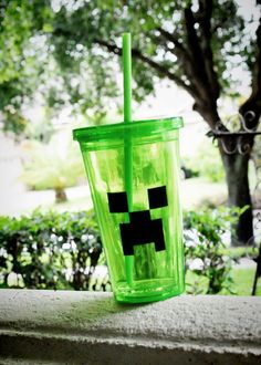 a green cup with a black face on it sitting in front of a tree and bushes