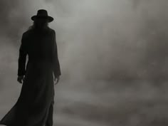 a man in a long black coat and hat standing on a foggy ground with his back to the camera