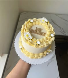 a hand holding a cake with yellow frosting and white flowers on the top, in front of a marble table