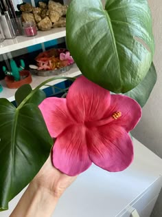 a person holding up a pink flower with green leaves in front of them on a table
